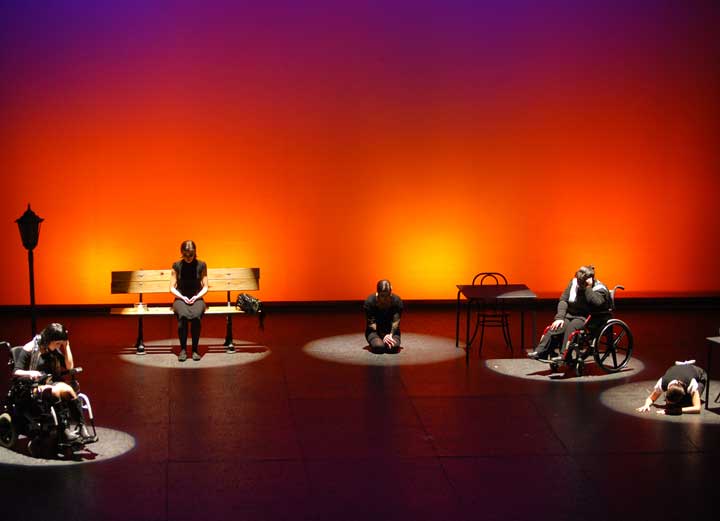 Foto de Maset de Frater en escena: tres personas sentadas en silla de ruedas, otra en un banco, y otra en el suelo, con una iluminación cálida en claroscuro